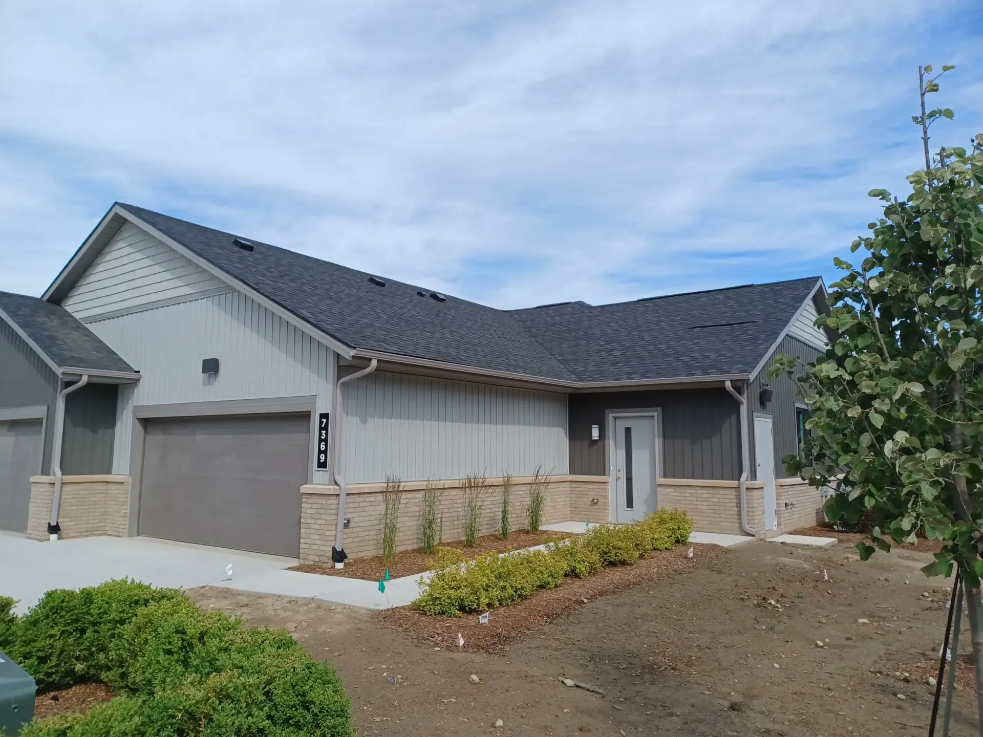 Gray house with a dark roof, a light gray garage door, and landscaping under a cloudy sky.