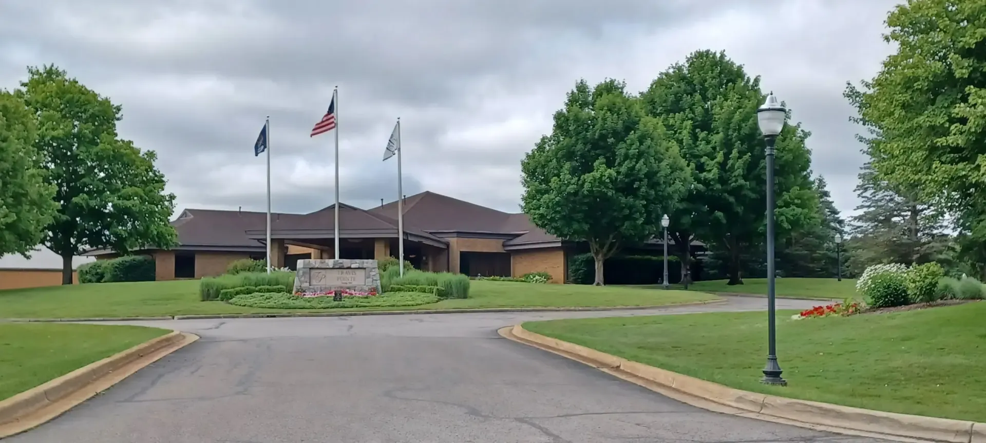 A building with flags in front of it, on a cloudy day. Green trees and grass with a paved driveway.