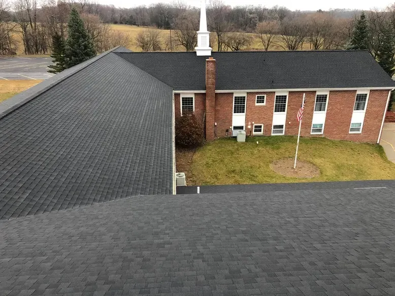 Church building with dark gray shingle roof, brick exterior, spire, and grassy lawn.