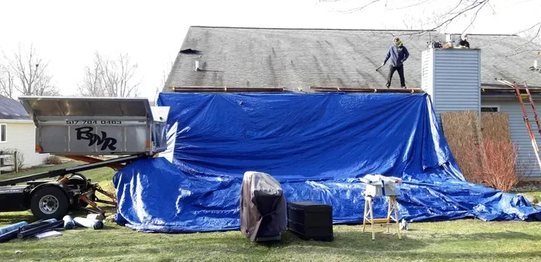 Man on roof with blue tarp covering house, equipment in yard.