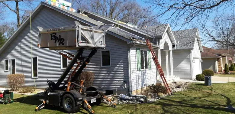 A roof is being worked on with a material lift. A ladder is propped against the house, and a truck is in front.