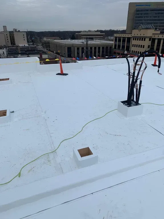 White flat rooftop with conduit, safety cones, and city buildings in the background under a cloudy sky.