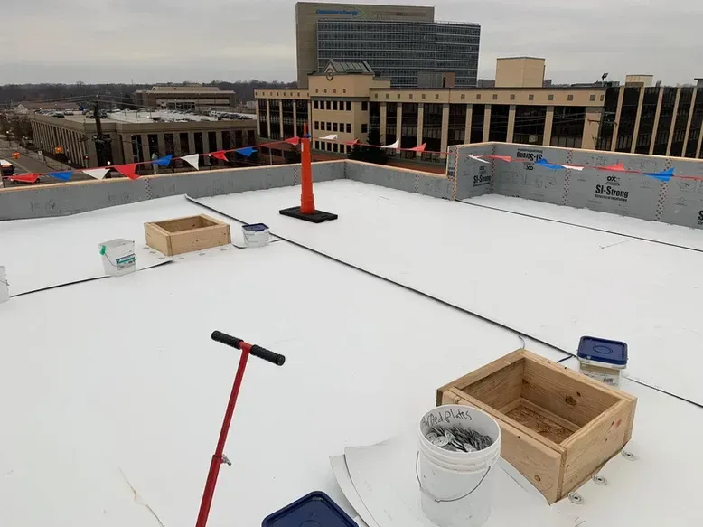Rooftop with construction materials: white surface, wooden boxes, buckets, red handle tool, urban background with buildings.