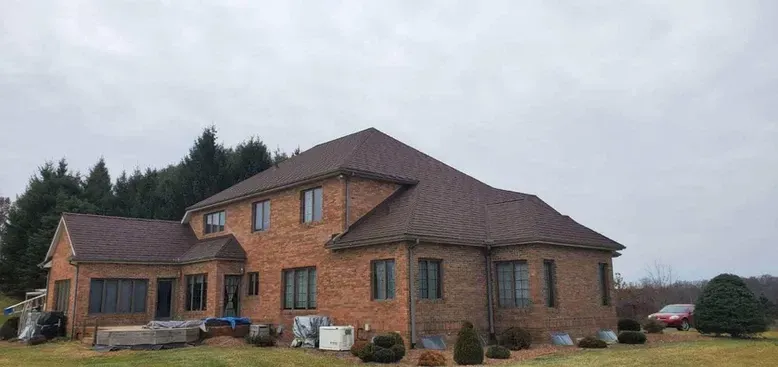 A large brick house with a brown roof stands on a grassy lawn under an overcast sky.