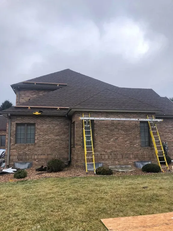 Brick house with roofers on ladders, cloudy day. Yellow ladders against brick wall, brown roof.