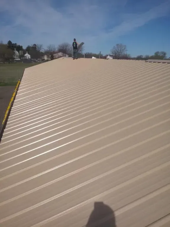 Person standing on a beige corrugated metal roof under a blue sky. Shadow visible.
