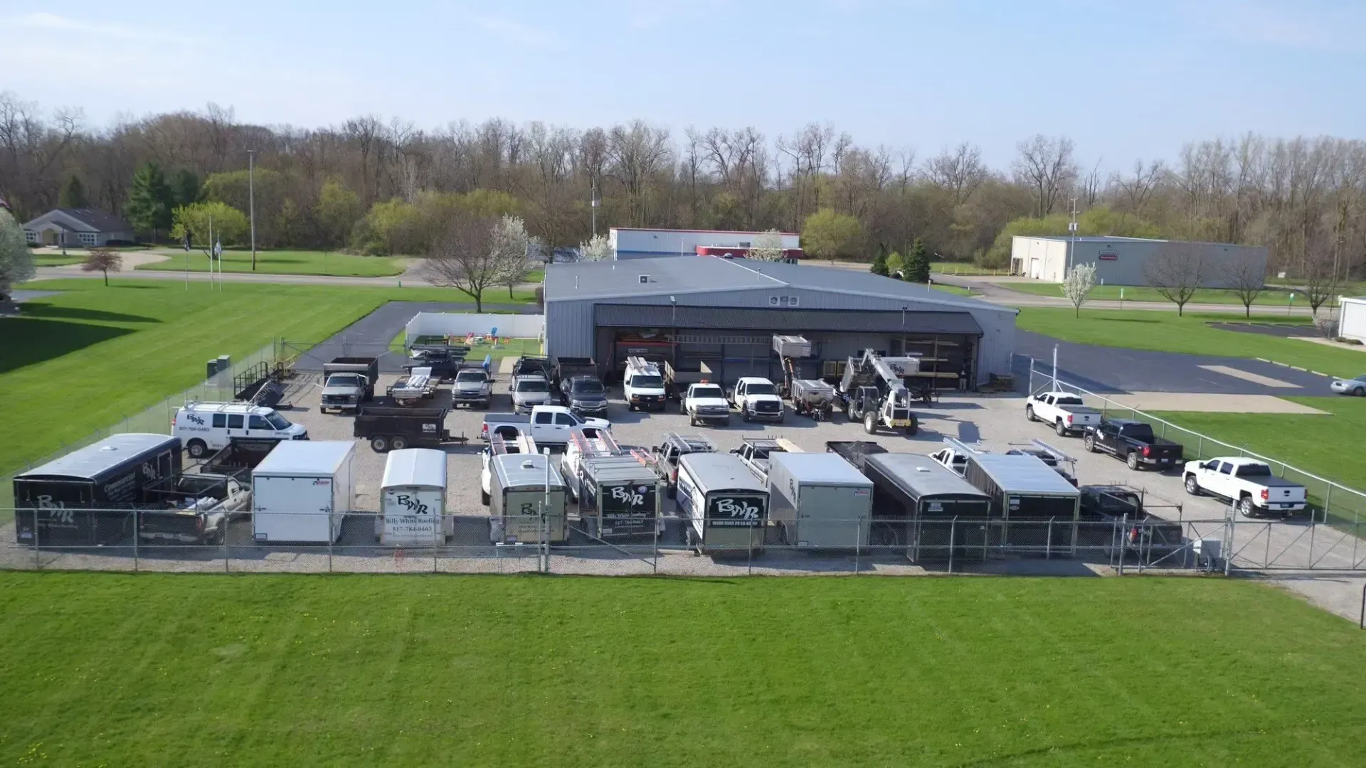 Vehicles and equipment parked in front of a gray building and grass. Fence surrounds the area.