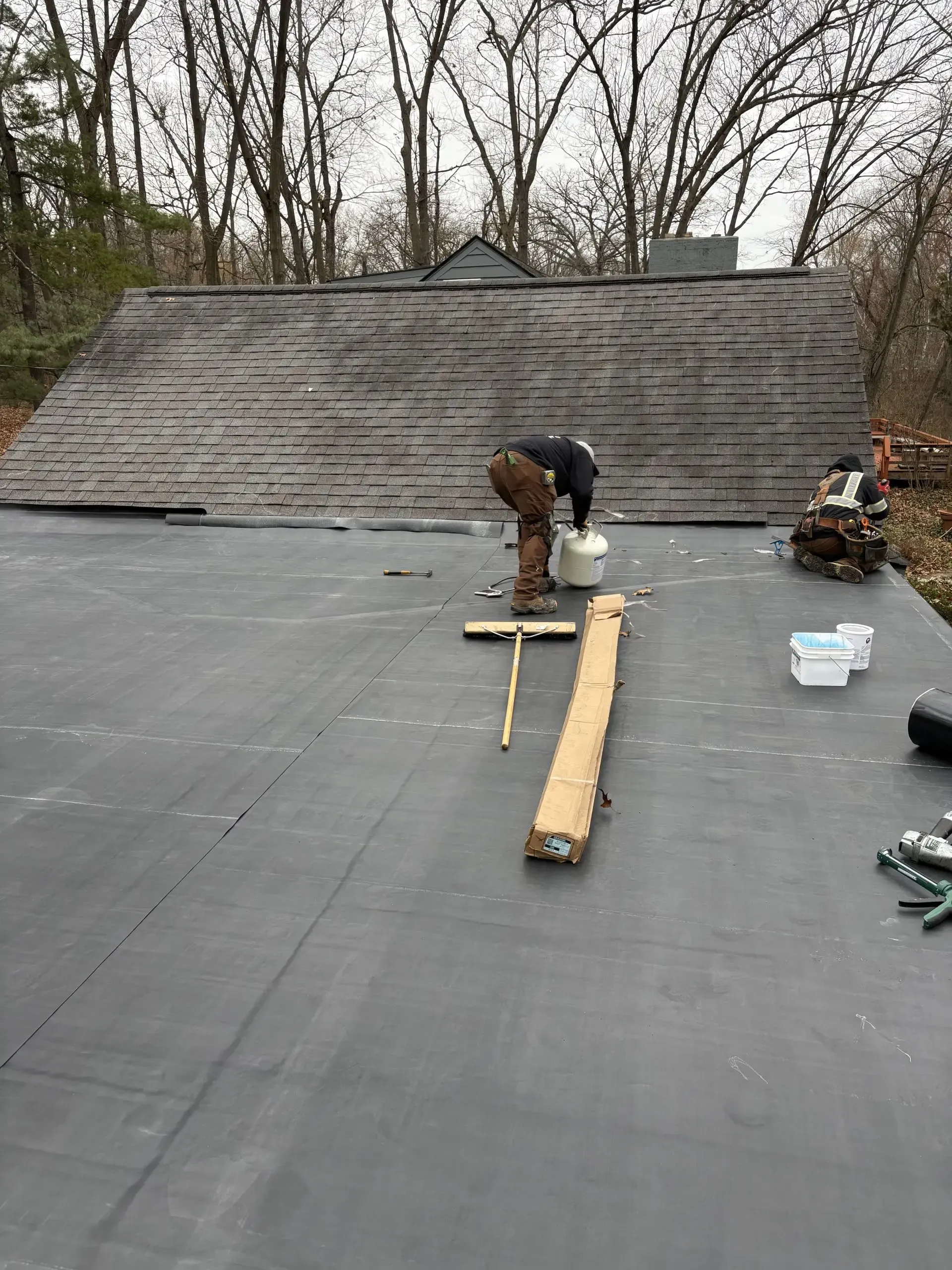Roofer on a black roof, working near wood and tools. Weathered, shingled roof visible in the background.