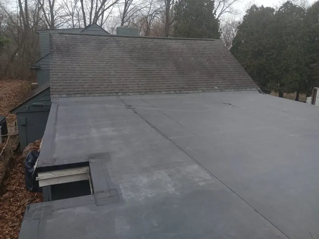 Dark gray flat roof of a building, with a smaller, peaked roof behind it; outdoor setting.