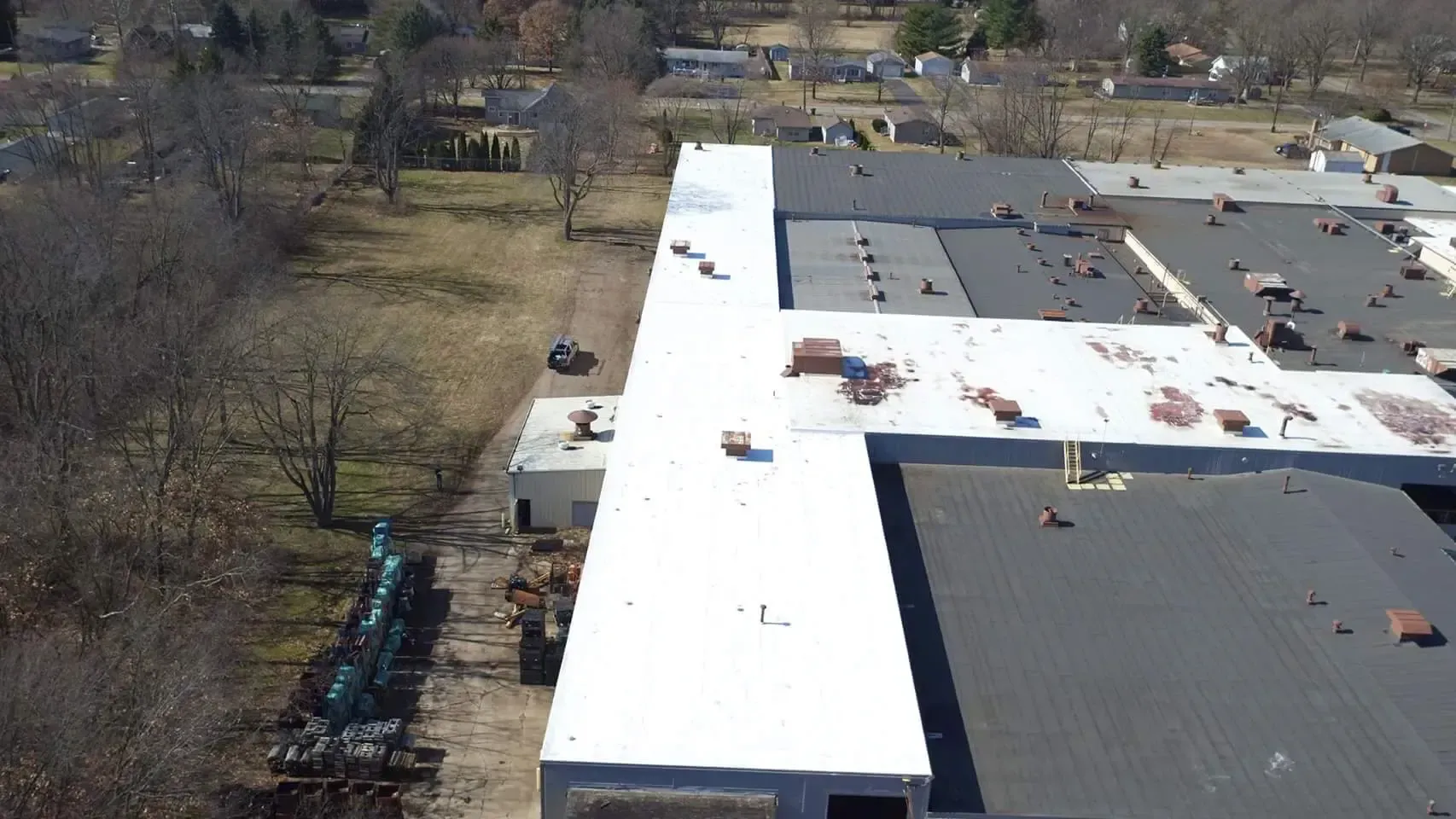 Aerial view of a large industrial building with a white roof and a dark roof. Trees and a field are in the background.