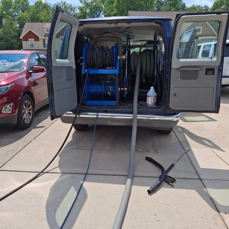 A cleaning van with open doors, hoses extended, parked on a driveway next to a car.