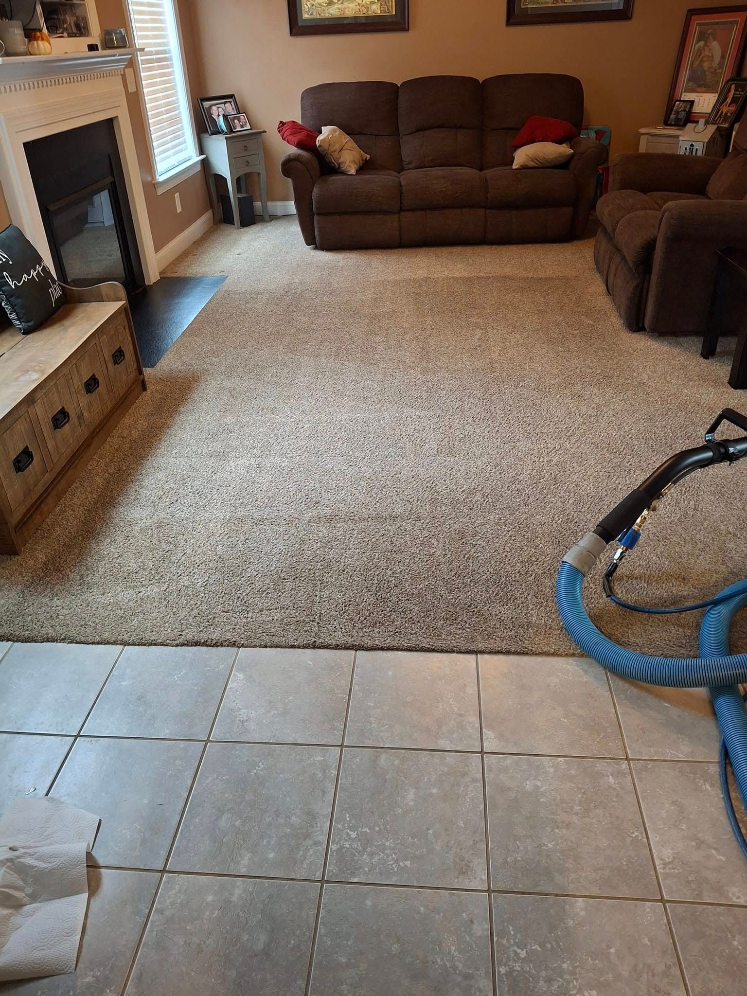 Carpet being cleaned with a blue hose; living room with brown couch and stone tile floor.