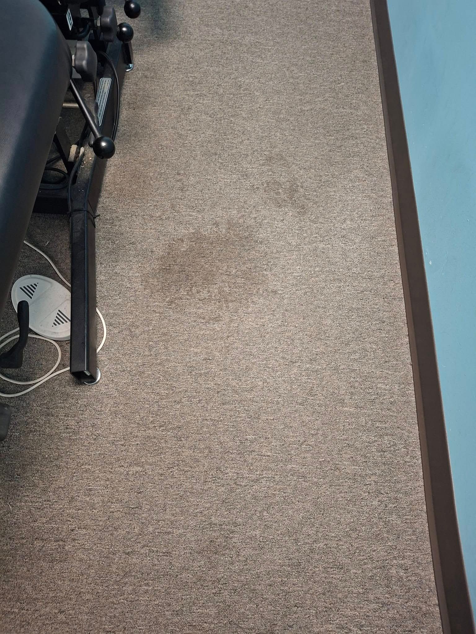 Brown carpet with dark stain, next to a black medical table leg and a light blue wall.