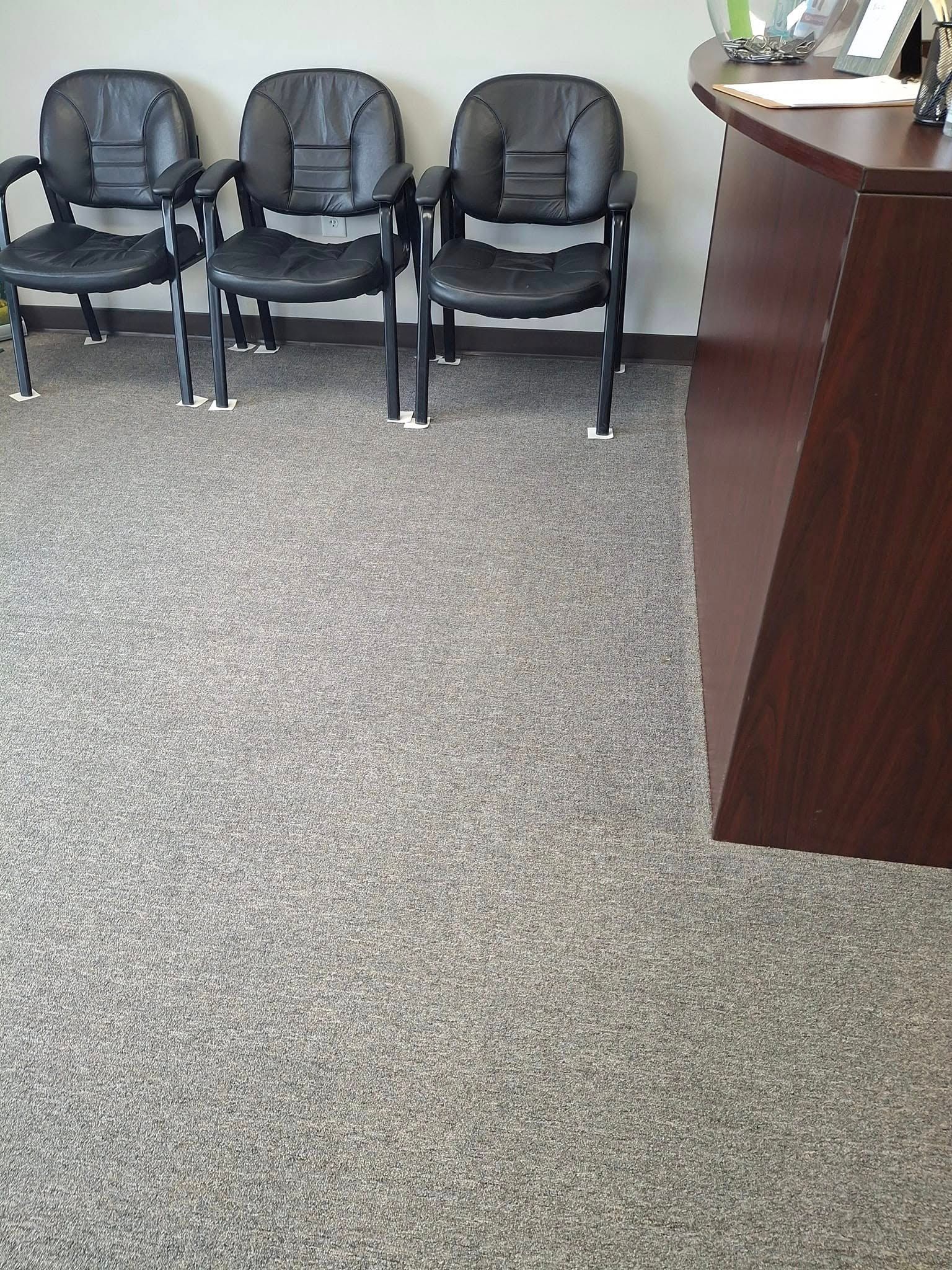 Three black chairs in a waiting area near a dark wood reception desk on speckled gray carpet.