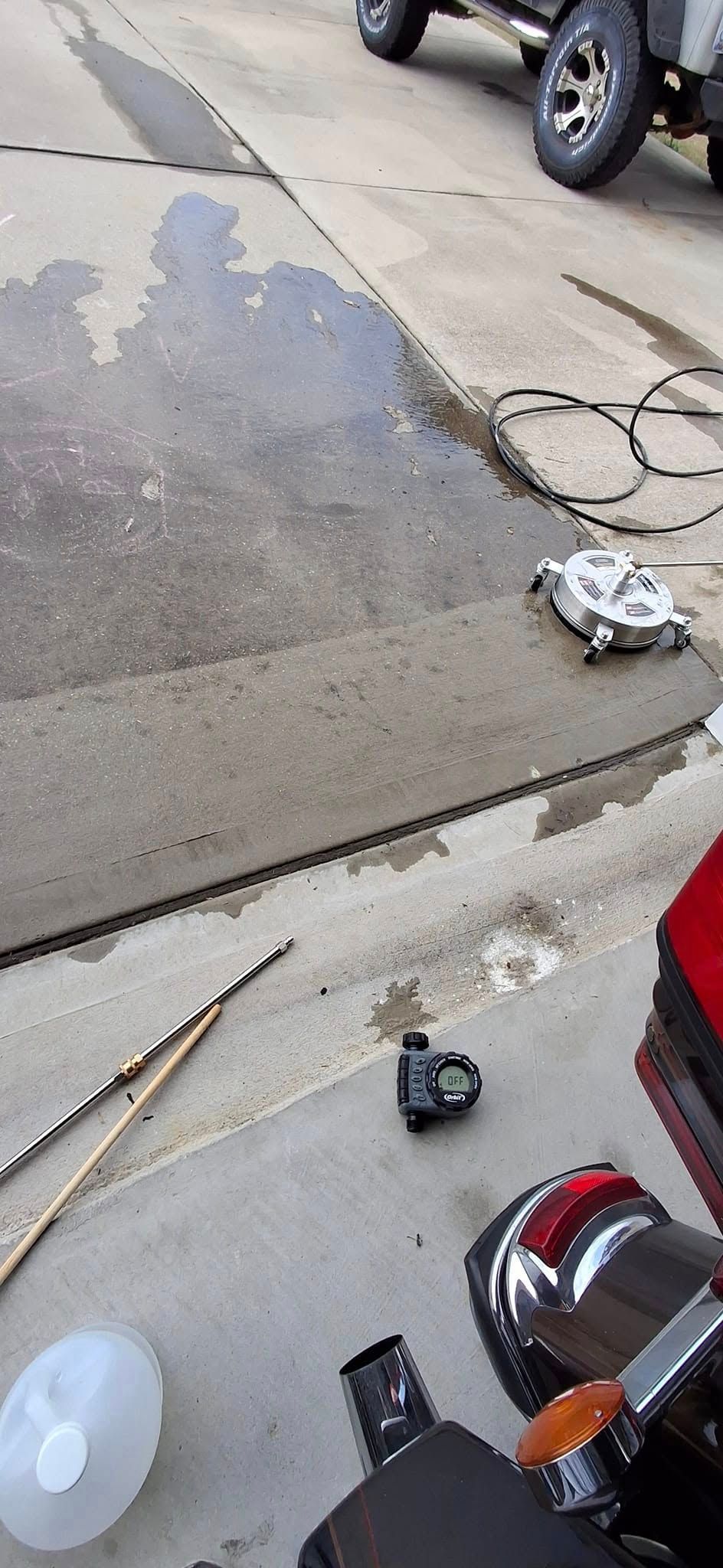 A motorcycle and car sit on a wet concrete surface. Tools and a jug are visible.