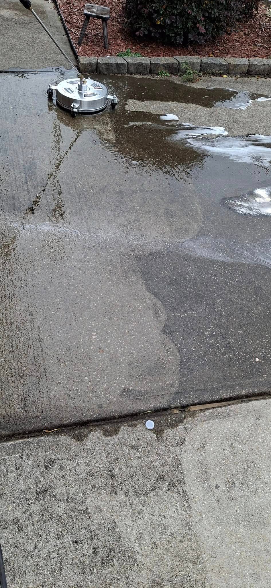 A concrete driveway being power washed with a circular surface cleaner, creating a large puddle near a bush.