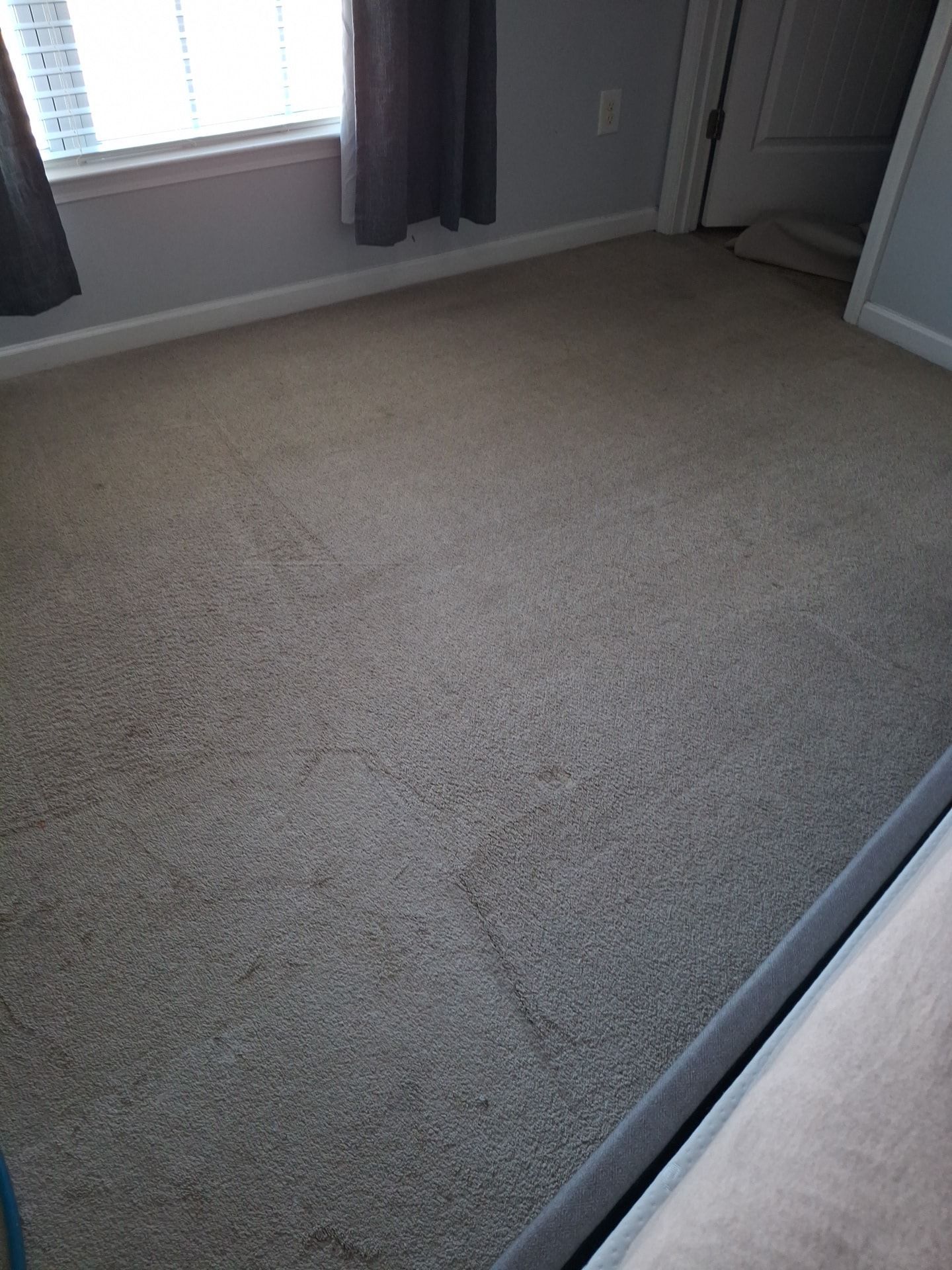 Beige carpeted bedroom with a window, curtains, and a partially visible bed frame.