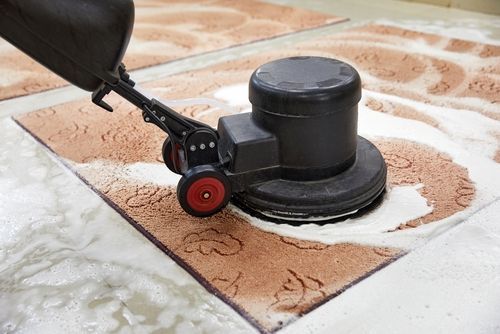 A machine scrubbing a brown rug, surrounded by soapy water, on a tiled floor.