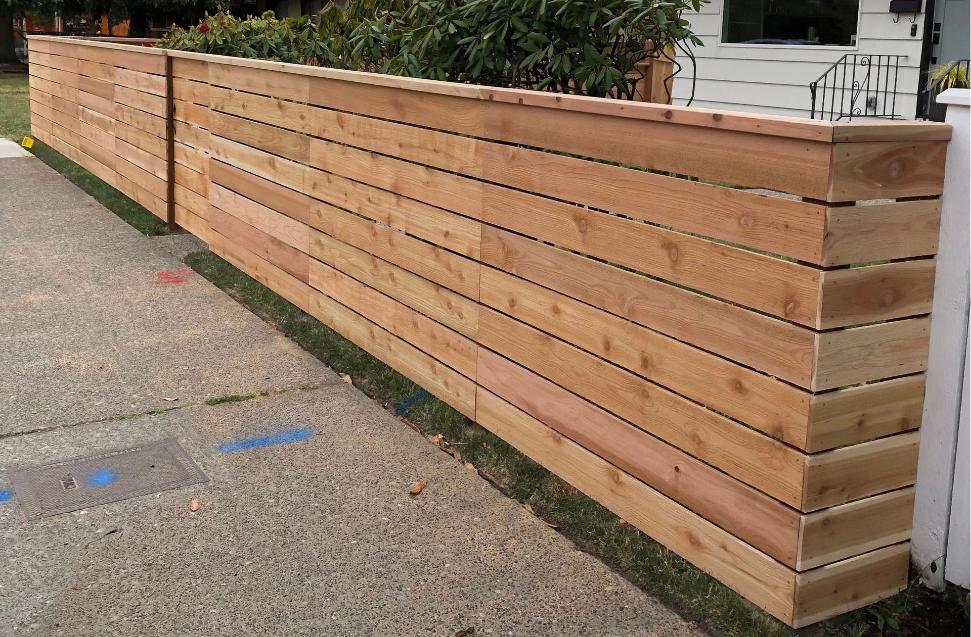 Wooden horizontal slat fence along a sidewalk, next to a house with grass.