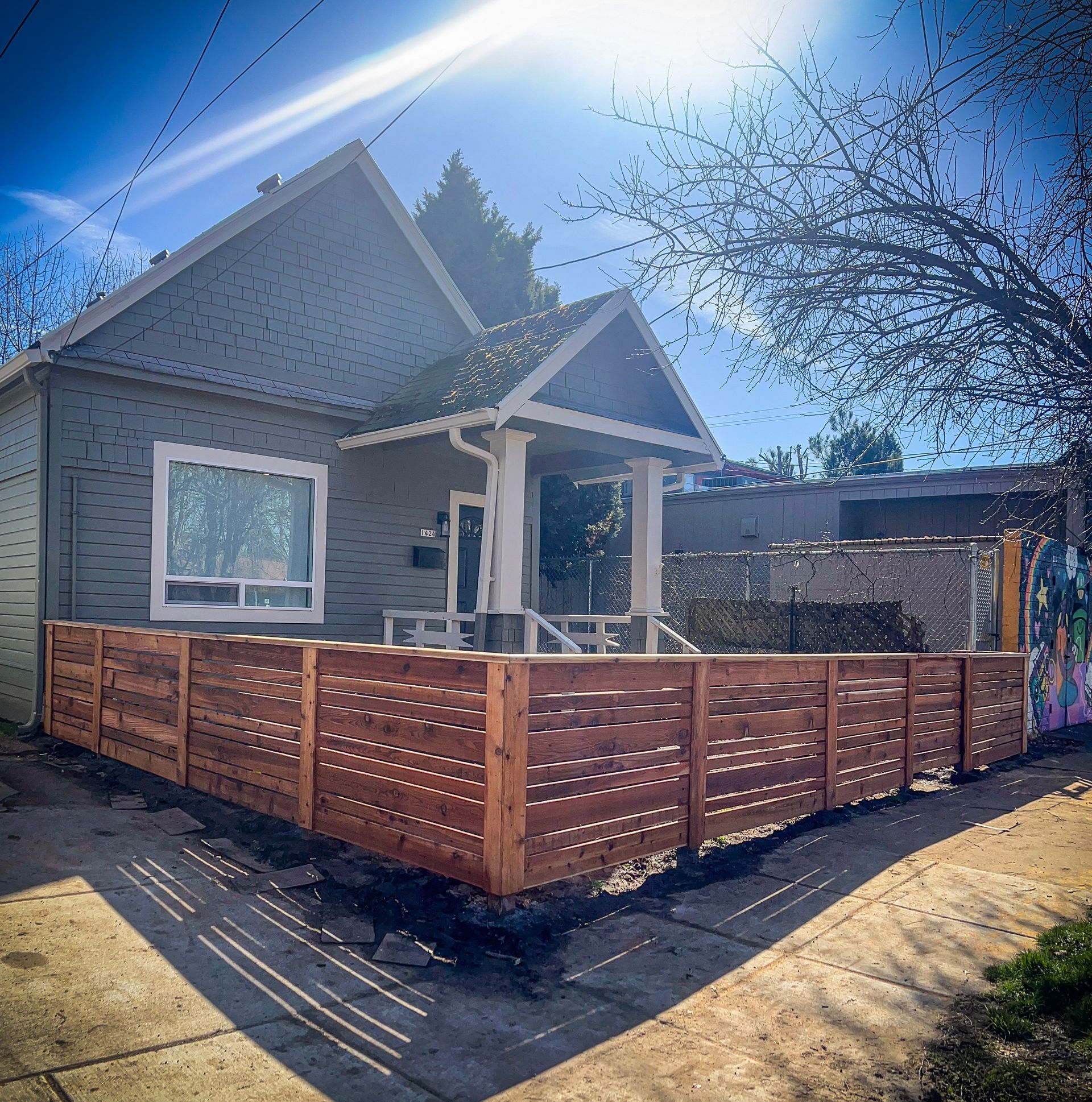 Gray house with a wooden fence in front, blue sky with bright sun, street view.