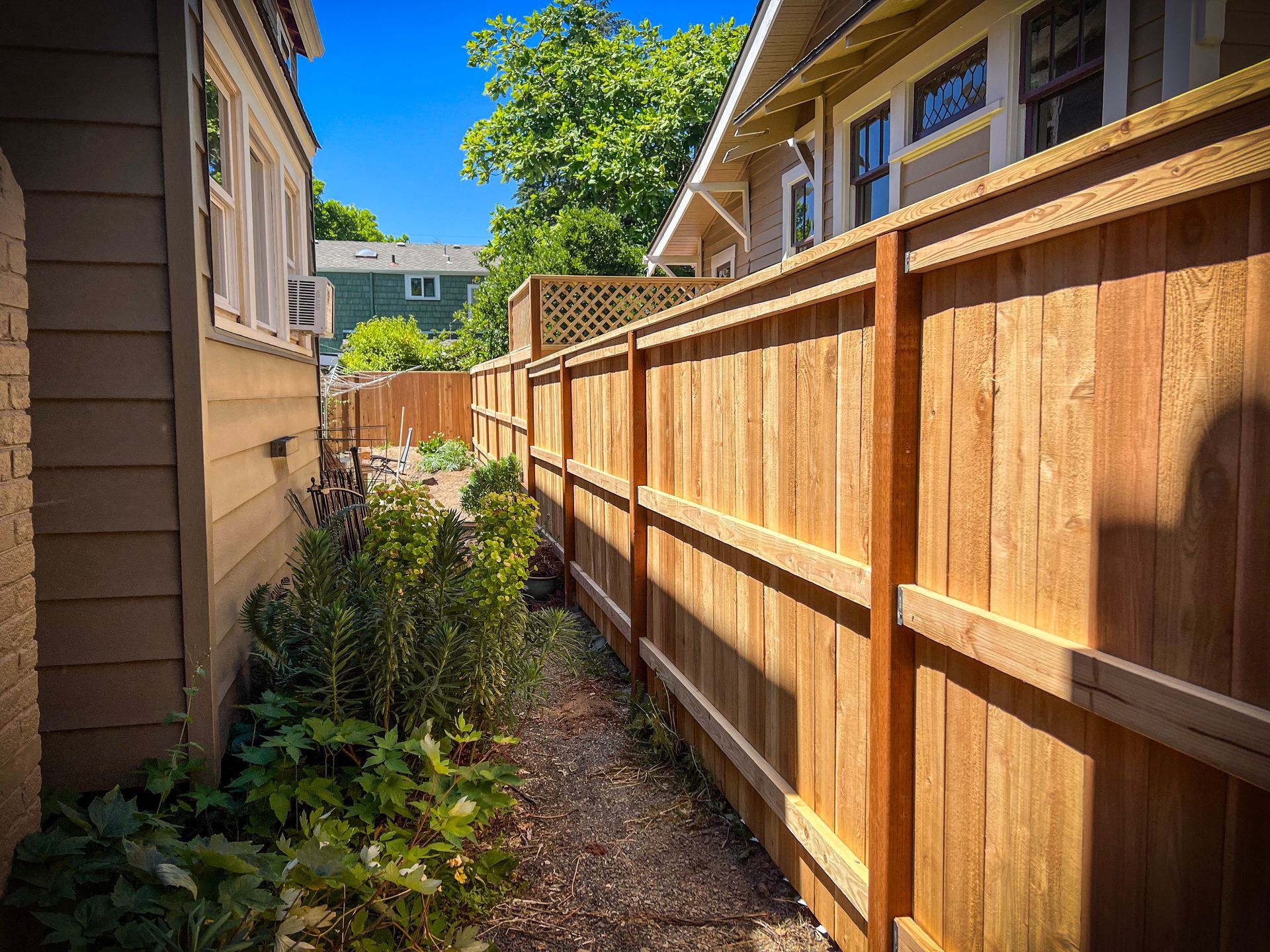 A wooden fence separates two buildings, with a narrow, shaded pathway running between them.
