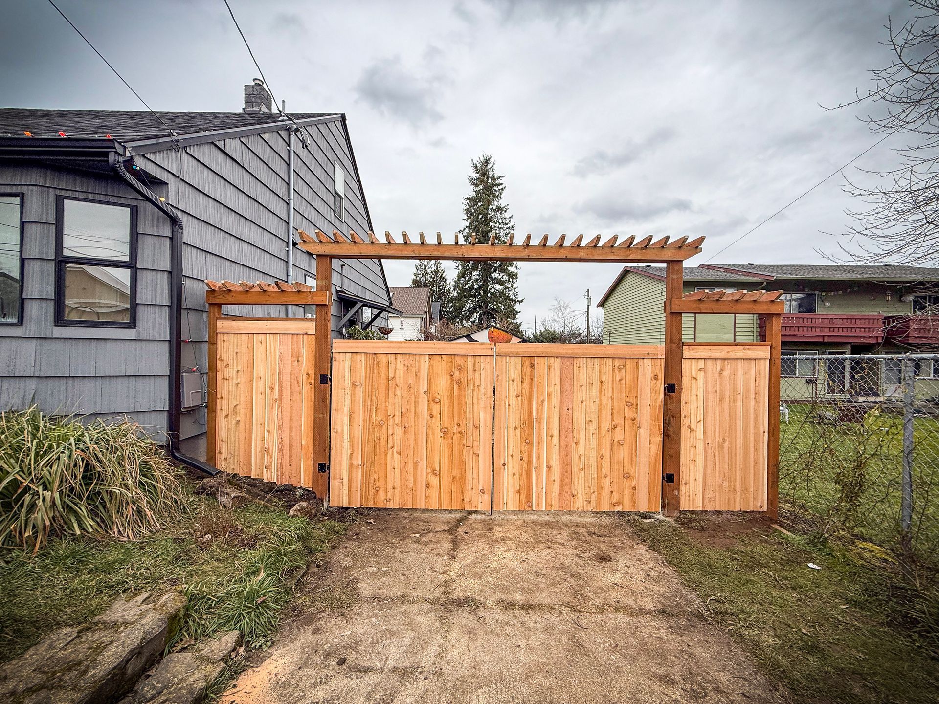 Wooden privacy gate with pergola detail, driveway. Brown fence in front of gray house.