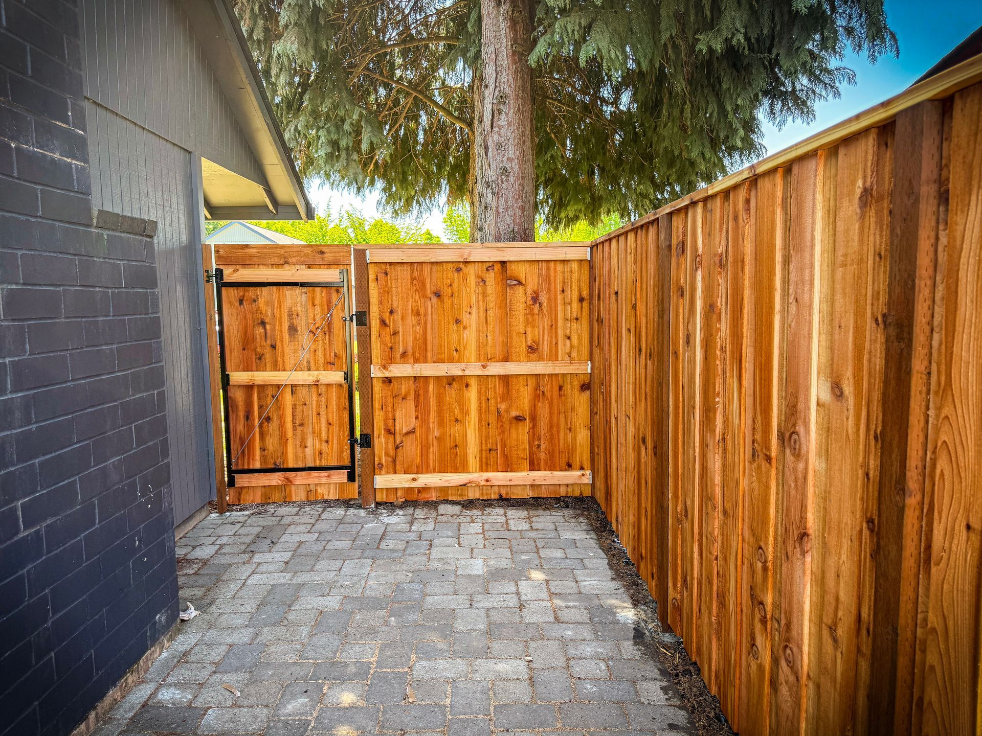 Wooden fence and gate enclose a brick-paved area next to a dark brick wall.