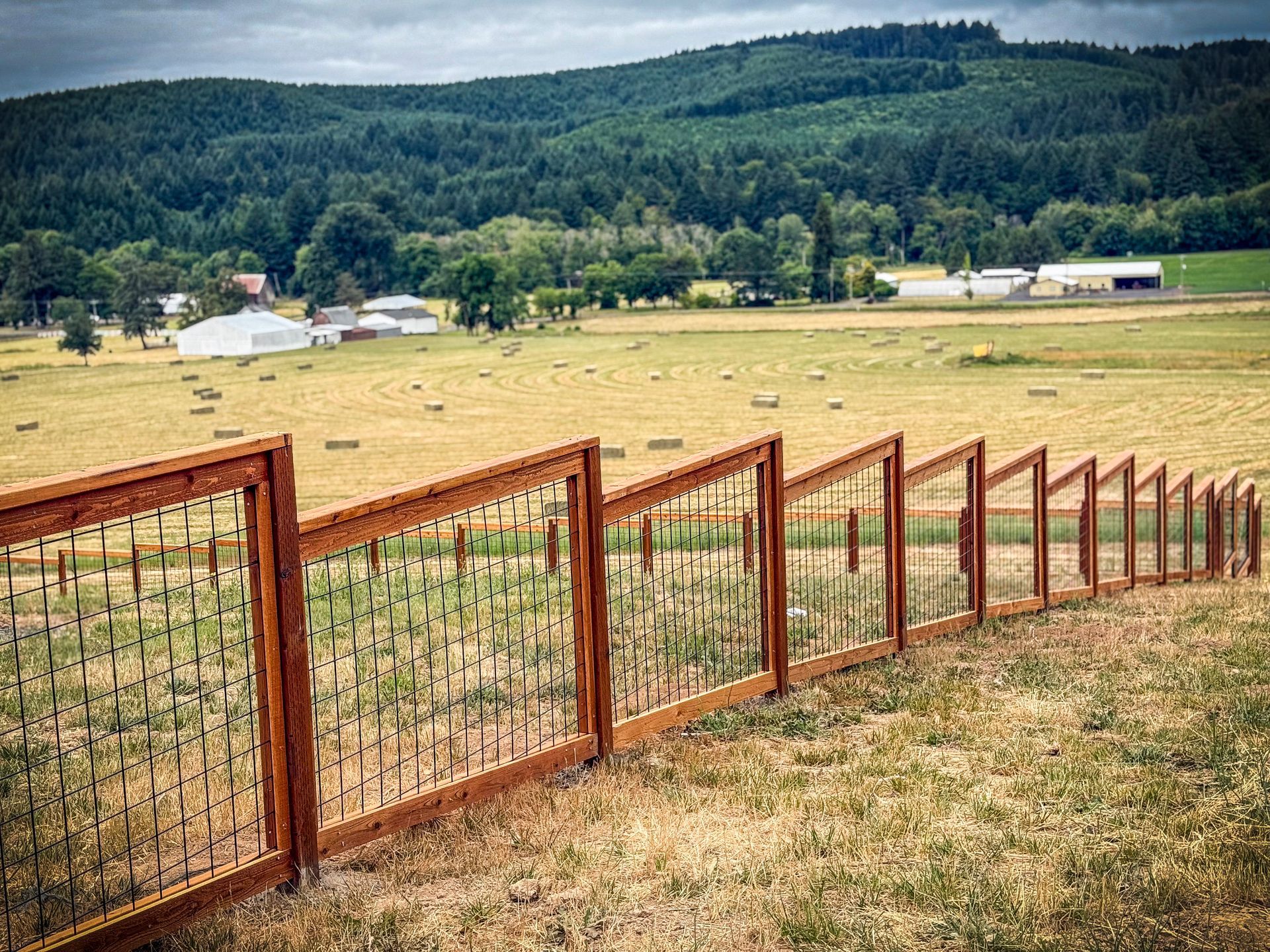 Wooden fence in a field with hay bales, buildings, and a forested hillside in the background.