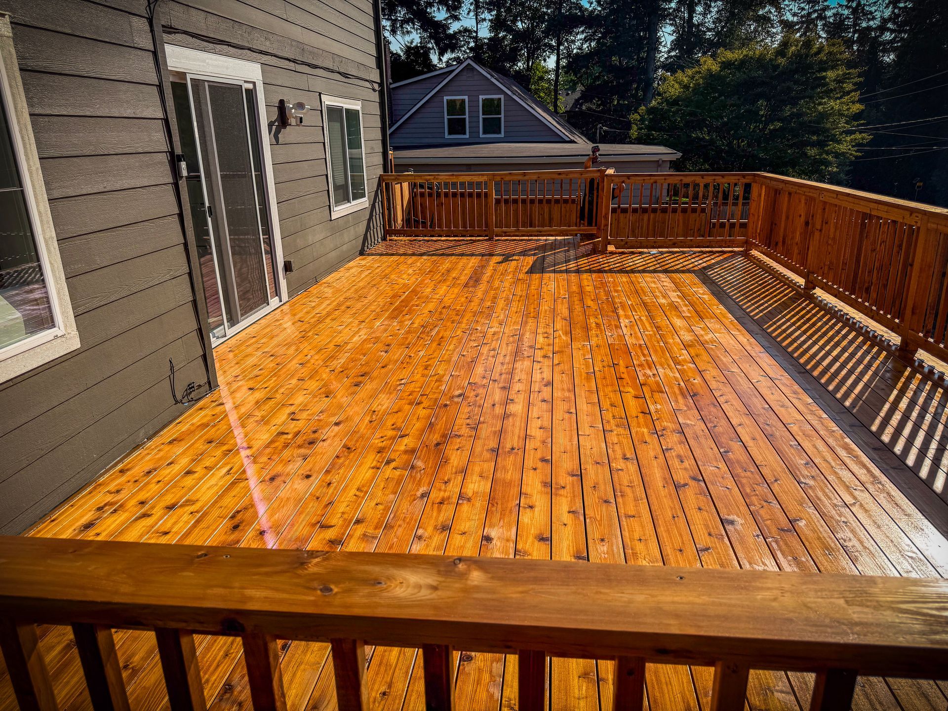 Wooden deck attached to a gray house, stained and glistening in sunlight.