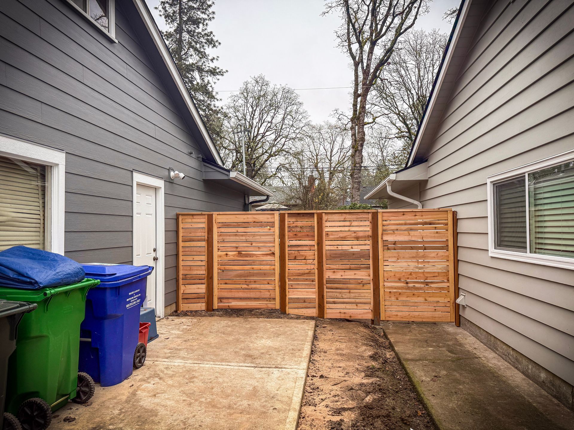 Wooden fence between two gray buildings, with trash cans and a muddy patch in front.