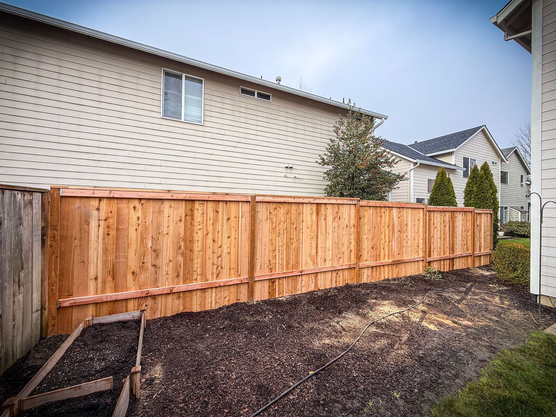 Wooden fence in a backyard; two-story beige house in background, dark mulch ground.