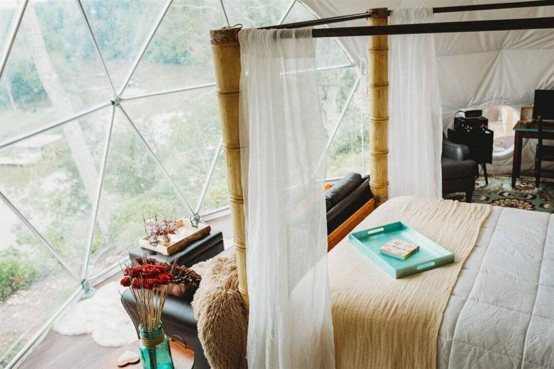 Bedroom inside a geodesic dome with bed, curtains, and a view of trees and water.