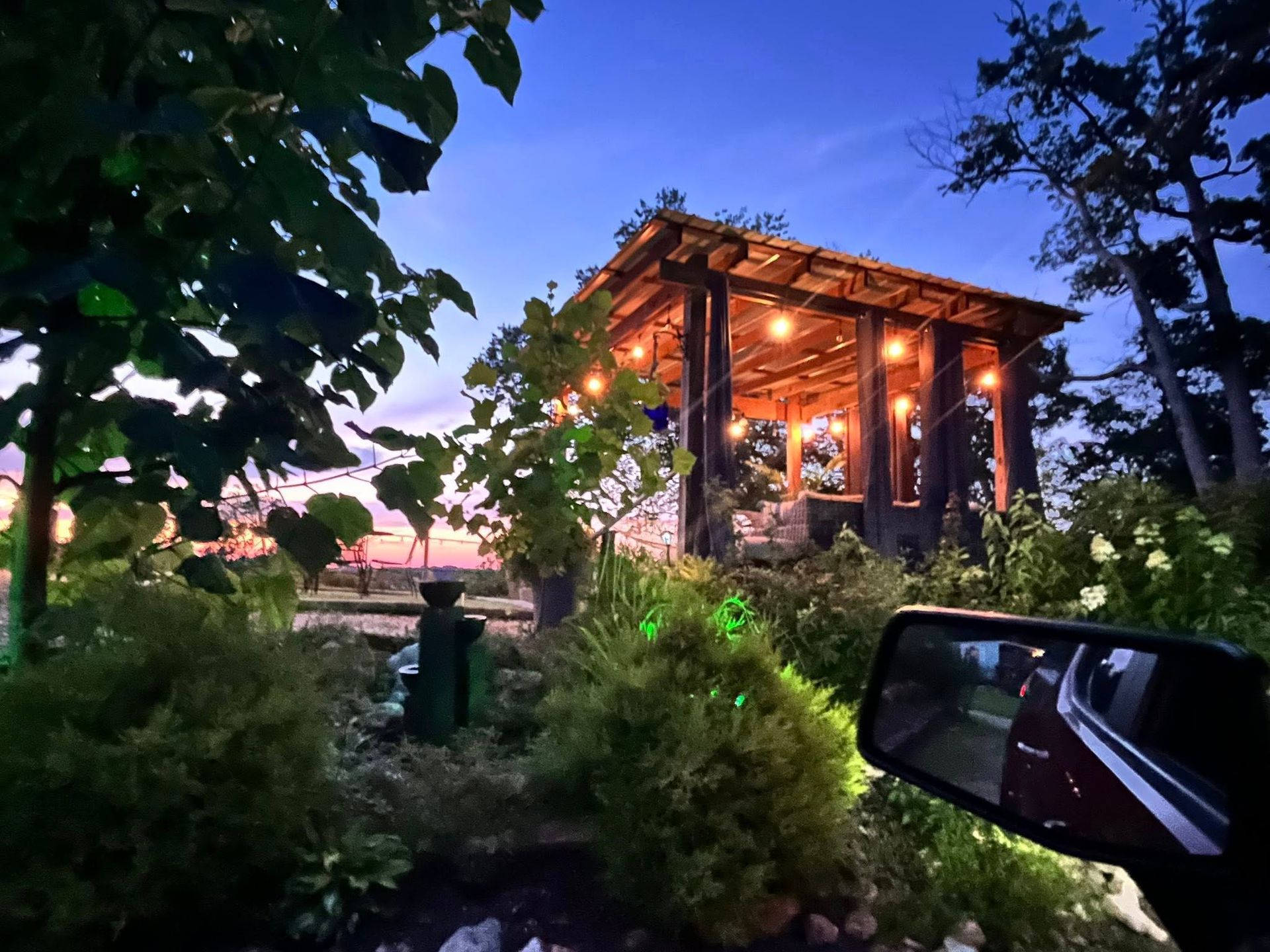 A rustic gazebo with string lights glows at dusk. Lush greenery surrounds the structure, visible from a car's side mirror.