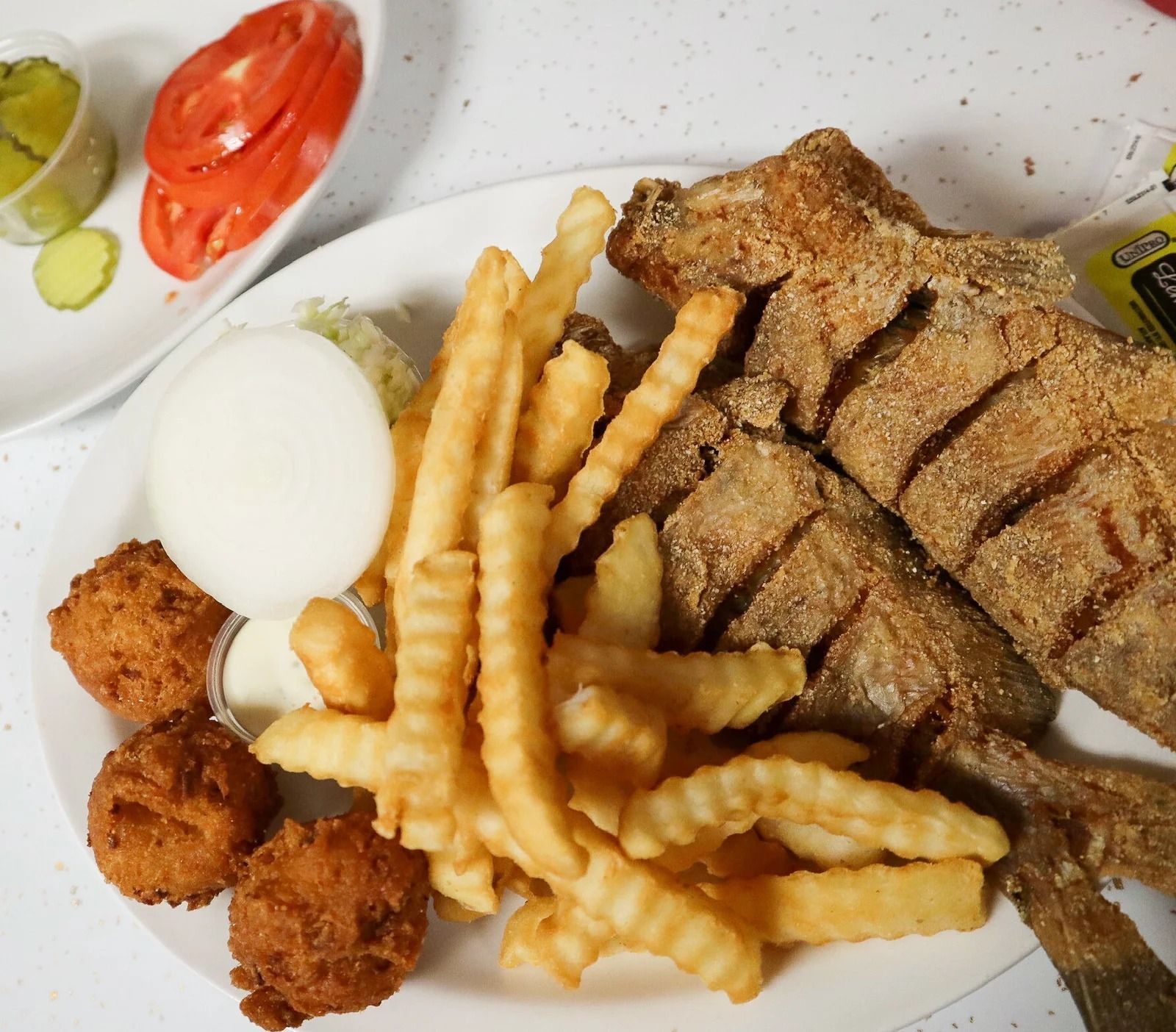 Fried fish plate with fries, hushpuppies, onion, coleslaw, pickles, and tomato.