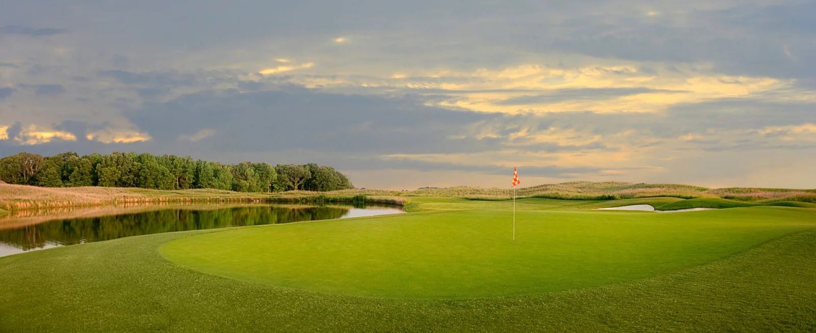 Golf green with a red flag, water feature, and trees under a cloudy sky.