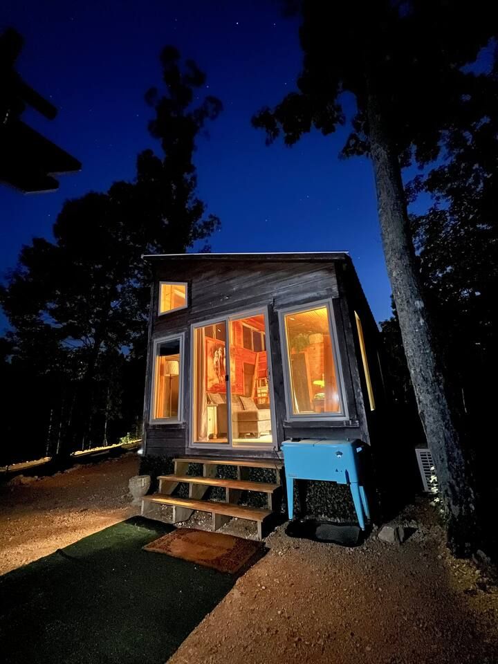 Tiny, illuminated cabin at dusk with open door and steps, surrounded by trees.