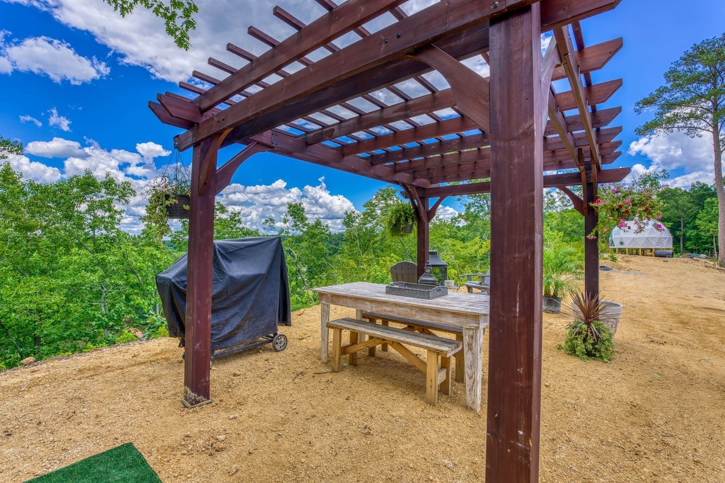 Pergola with picnic table, grill, and hanging plants outdoors; sunny day.