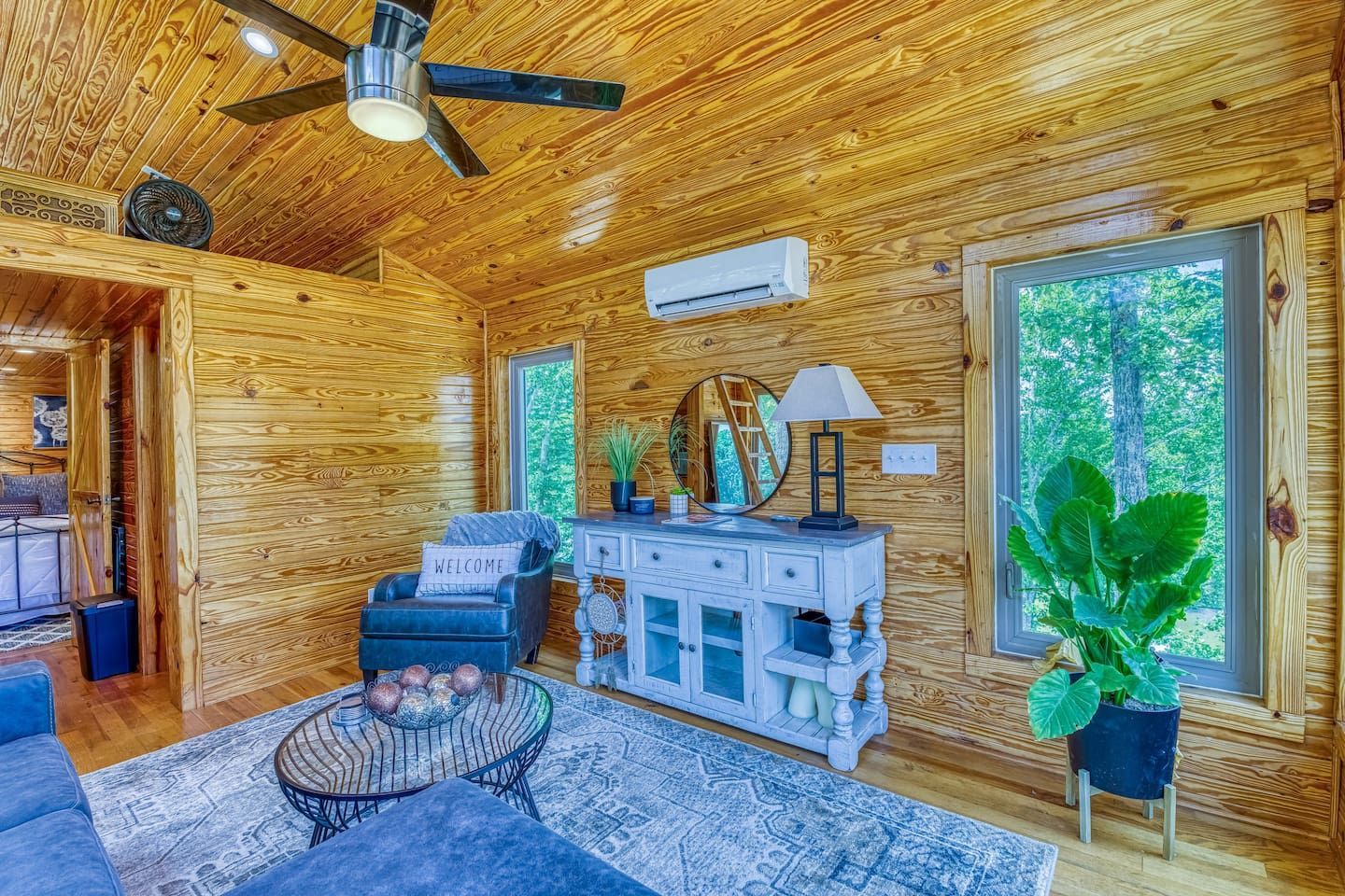 Cozy living room with wood paneling, blue armchair, white sideboard, rug, large window, and indoor plant.