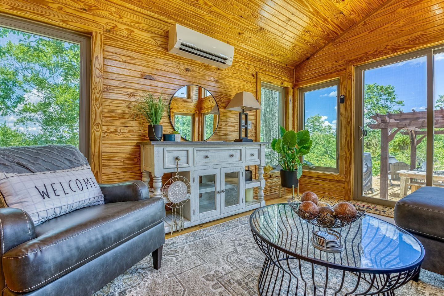 Cozy sunroom with wood-paneled walls, leather seating, and views of trees through large windows.