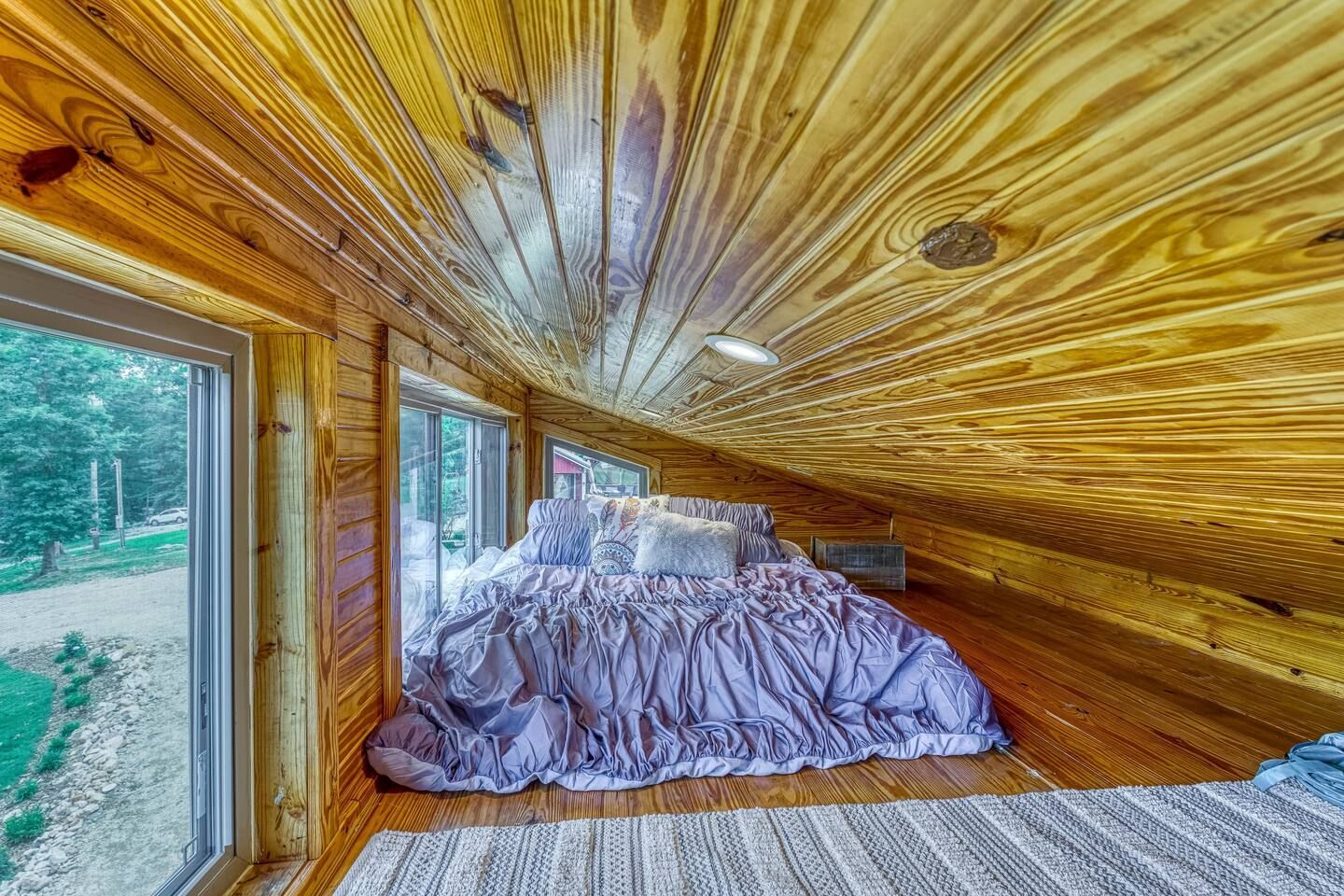 Loft bedroom with wooden walls and ceiling, large bed with lavender bedding, and windows.