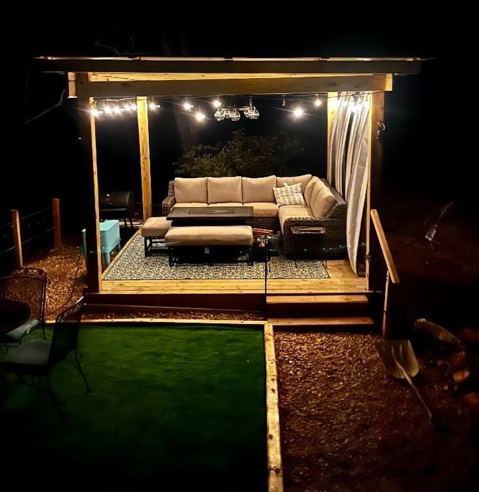 Lit wooden gazebo with couch, rug, and string lights at night.