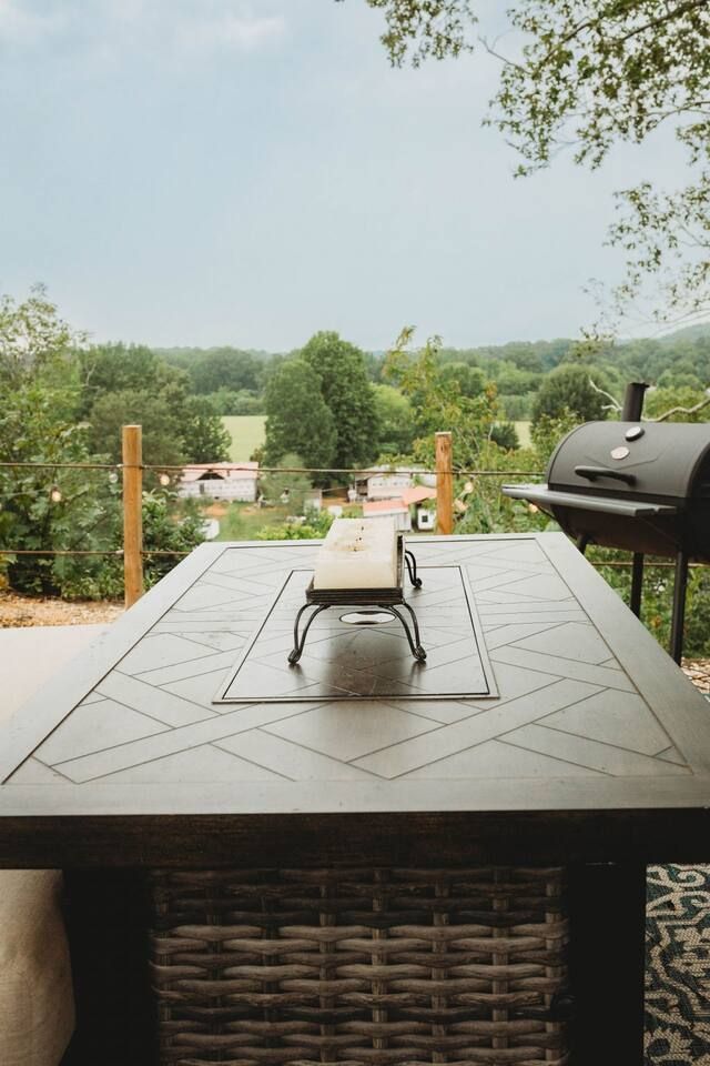Outdoor dining table with fire pit, grill, and distant view of trees and fields.