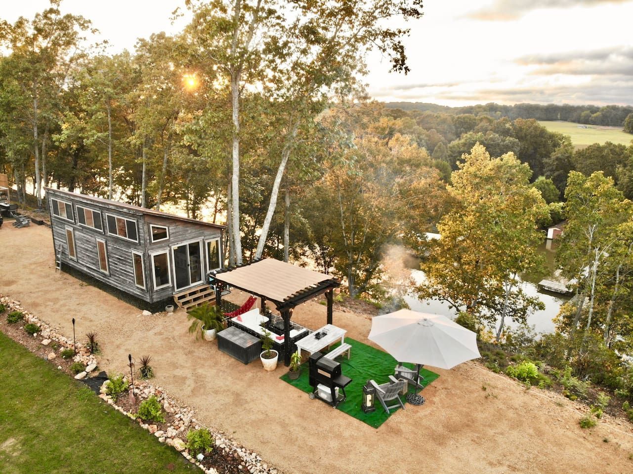 Tiny house near water with outdoor dining and a grill. Trees and fields in the background.