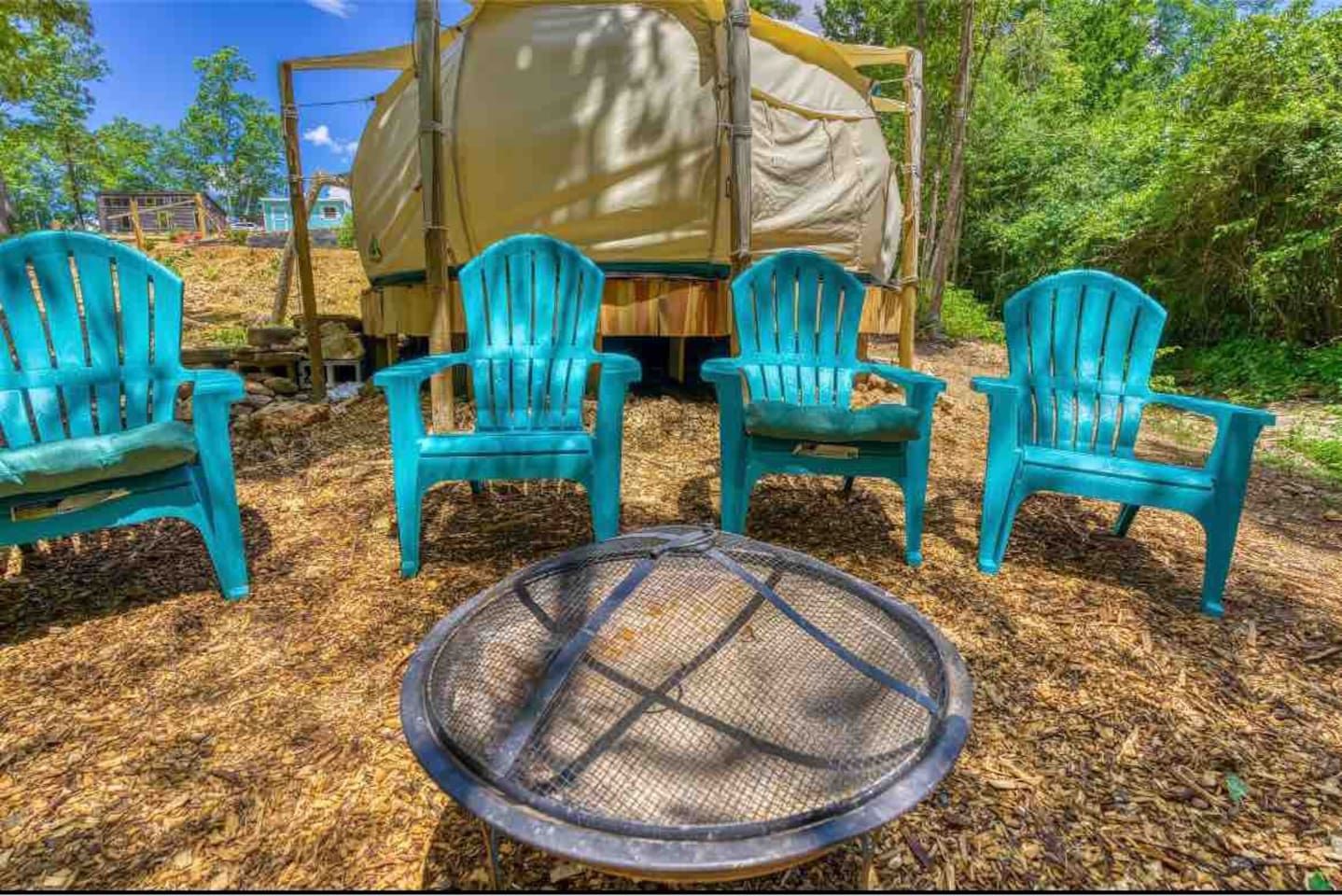Four teal chairs around a fire pit, with a yurt in the background, set in a wooded area.