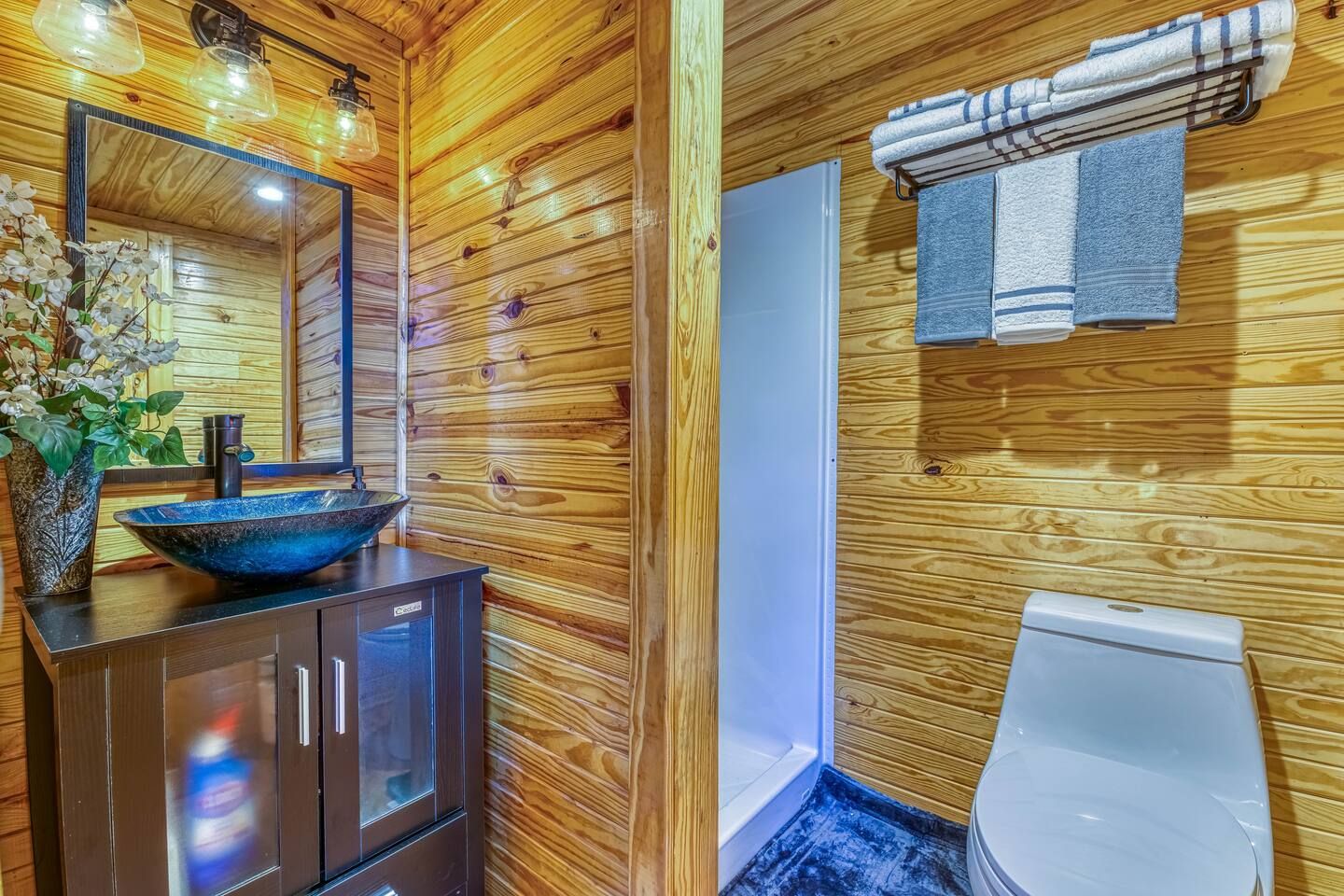 Bathroom with wood-paneled walls, a dark vanity with blue glass sink, a toilet, and towels hanging on a rack.