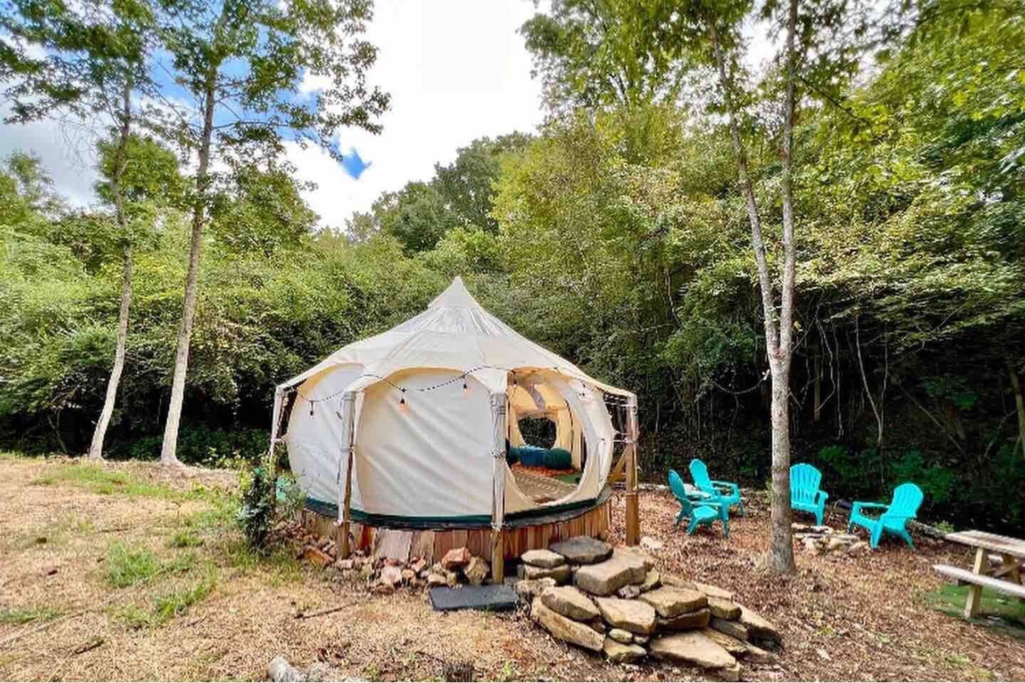 A glamping tent sits in a wooded area with blue Adirondack chairs nearby.