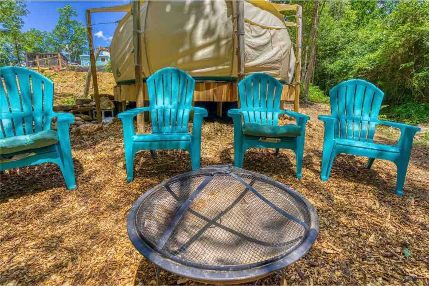 Four turquoise Adirondack chairs around a fire pit in a wooded area; yurt in the background.