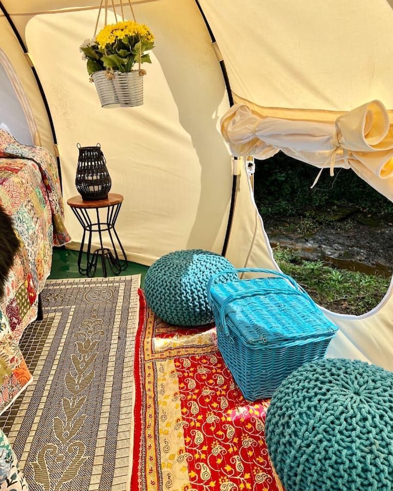 Cozy tent interior with rugs, poufs, basket, flowers, and a small table with a decorative item.