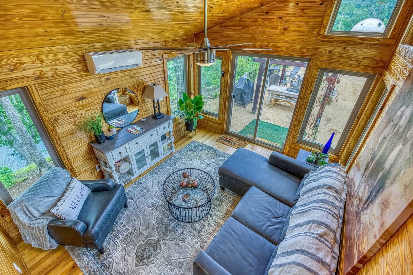 Cozy cabin living room with wooden walls, grey sofa, round rug, and a sliding glass door to a deck.