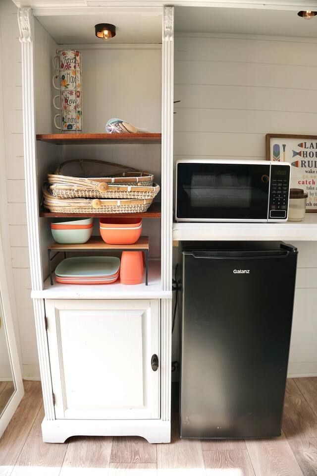 White cabinet with shelves holding dishes. A microwave and mini-fridge sit adjacent.